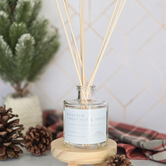 A reed diffuser bottle with natural wooden reeds, displayed on a small wooden stand. The bottle has a white label reading 'ROASTED CHESTNUT & SPICE'.