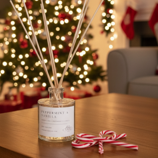 A peppermint and vanilla reed diffuser bottle with wooden reeds sits on a wooden table in a cozy living room, with a blurred Christmas tree and fireplace in the background.