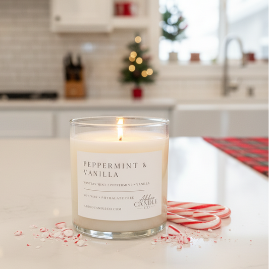A lit jar candle labeled 'PEPPERMINT & VANILLA' sits on a white kitchen counter with candy cane pieces scattered around it. The background shows a softly blurred holiday kitchen with lights and decorations.