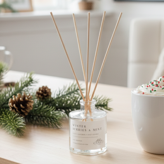 A clear glass reed diffuser bottle labeled 'Winter Vanilla & Mint' with four natural reeds, styled on a light wood table next to a white mug filled with whipped cream and sprinkles, with holiday greenery and a softly blurred Christmas tree in the background.