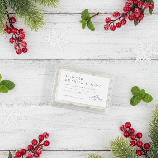 A wax melt package labeled 'Winter Berries & Mint' by Aroma Wicks sits on a white wood surface, surrounded by festive holiday props including pine branches, red berries, mint leaves, and white snowflake decorations.